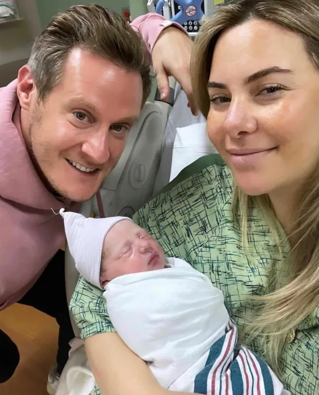 A smiling couple in a hospital with their newborn baby wrapped in a white blanket.
