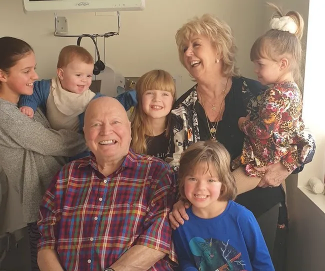 A smiling elderly man and woman with five children gathered around them, in a cozy indoor setting.