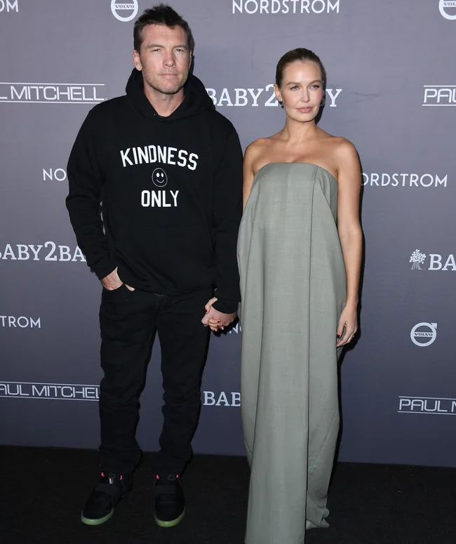 Couple on red carpet, man wears "Kindness Only" hoodie, woman in strapless dress, Baby2Baby event backdrop.