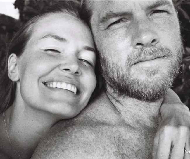 A smiling woman and a man with a beard pose closely together in a black-and-white photo.