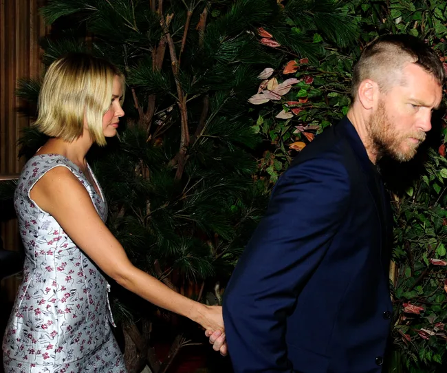 Two people walking, holding hands, near foliage. The woman wears a floral dress, and the man wears a dark blue blazer.
