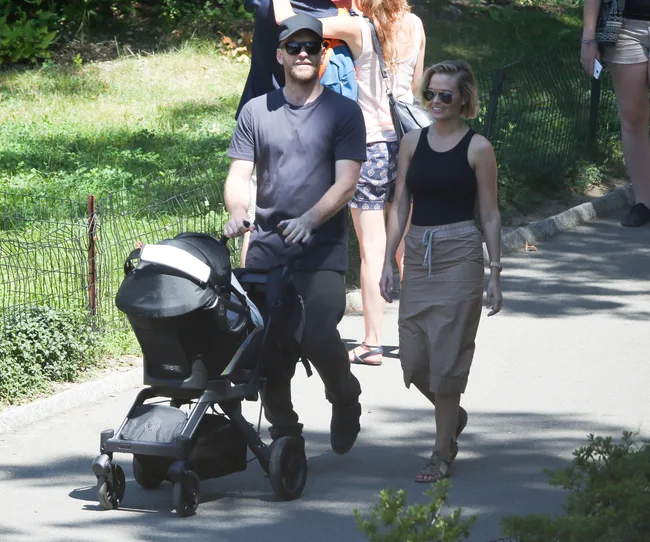 Man pushing a stroller and a woman walking beside him in a park on a sunny day, surrounded by greenery and other people.