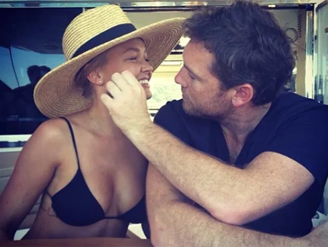 A couple sitting closely on a boat, the woman wearing a wide-brimmed hat, and the man gently touching her face.