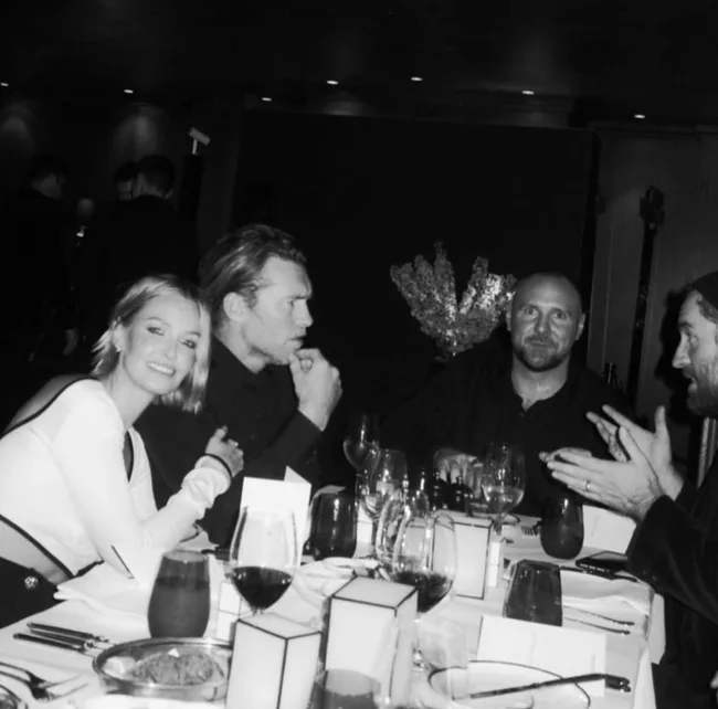 Four people in black and white at a dinner table, engaged in conversation with wine glasses and table settings.