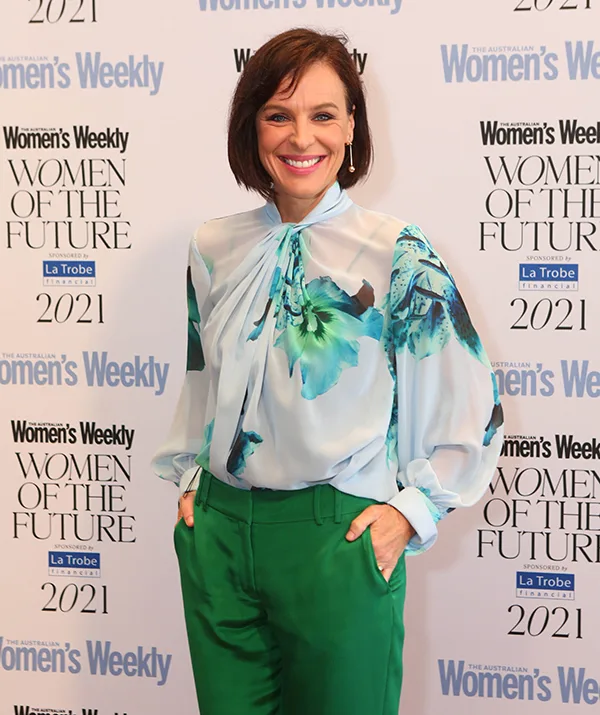 Woman in floral blouse and green pants at Women's Weekly "Women of the Future" event backdrop, smiling with hands in pockets.