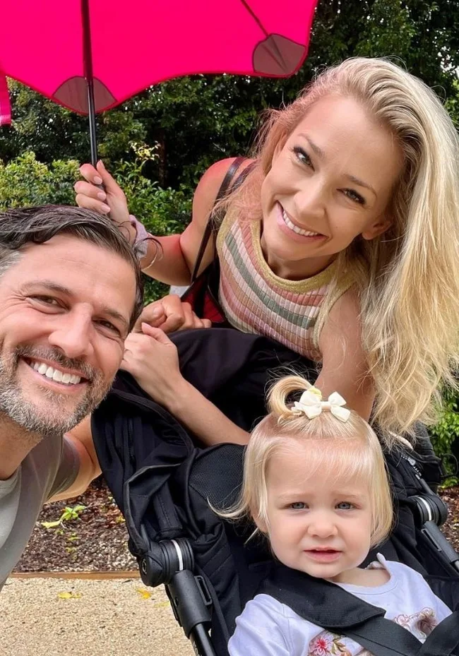 Family selfie under a pink umbrella with a smiling man, woman, and a child in a stroller.