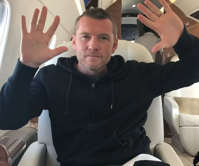 Man in a black hoodie sitting in an airplane, raising both hands with open palms, facing the camera.