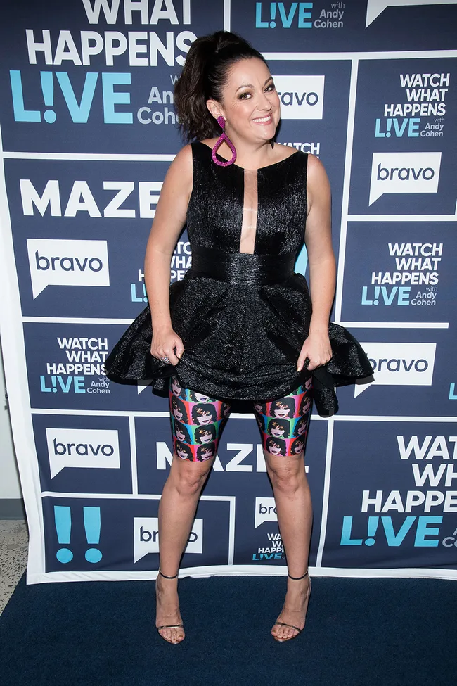 A woman in a black dress and colorful shorts poses at a "Watch What Happens Live" event.