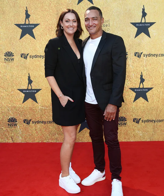 A man and woman posing on the red carpet with a "Hamilton" backdrop, both in black attire and white sneakers.