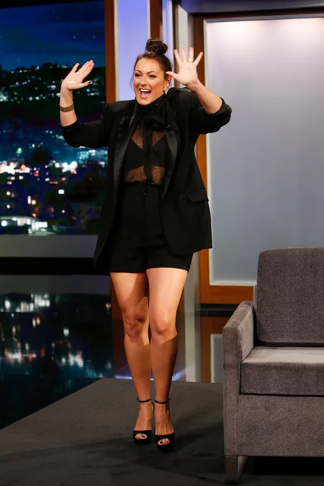 Person in a dark outfit excitedly waving hands on a talk show set with a nighttime cityscape background.