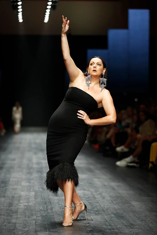 Model in a black dress and large earrings posing confidently on the runway.