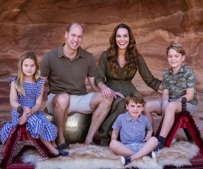 A family of five posing on a rug against a rocky background, all smiling warmly.