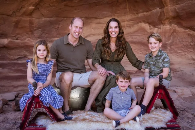 Royal family sitting together, smiling in casual outfits against a rocky background, with children on decorative rugs.
