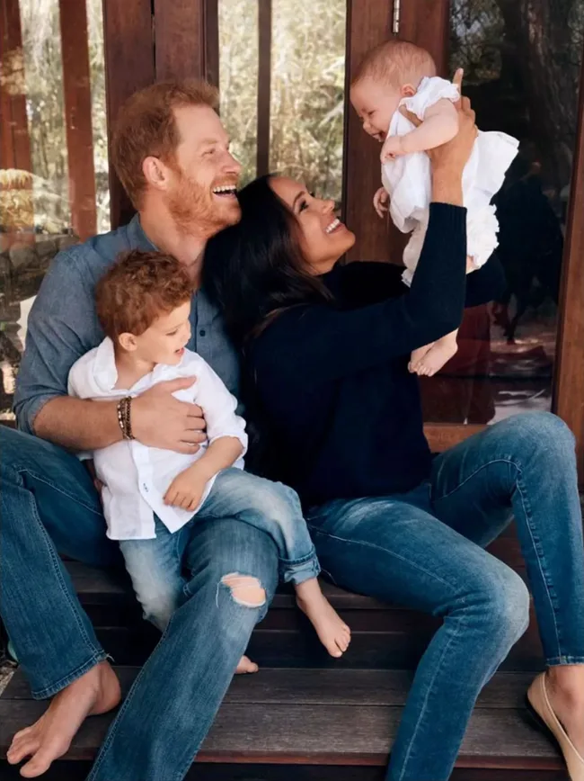 A family of four sits together on a porch, the parents happily playing with their children, one toddler and one baby.