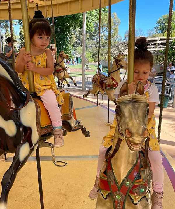 Two young girls in yellow outfits riding a merry-go-round horse in a park with trees in the background.