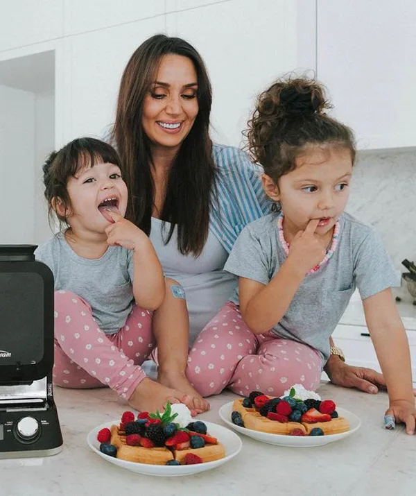 A woman and two young girls in pajamas sit at a kitchen counter with fruit-topped waffles, smiling and playful.