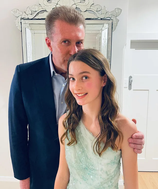 Father in a suit stands behind his daughter in a mint dress, both smiling with a decorative mirror behind them.