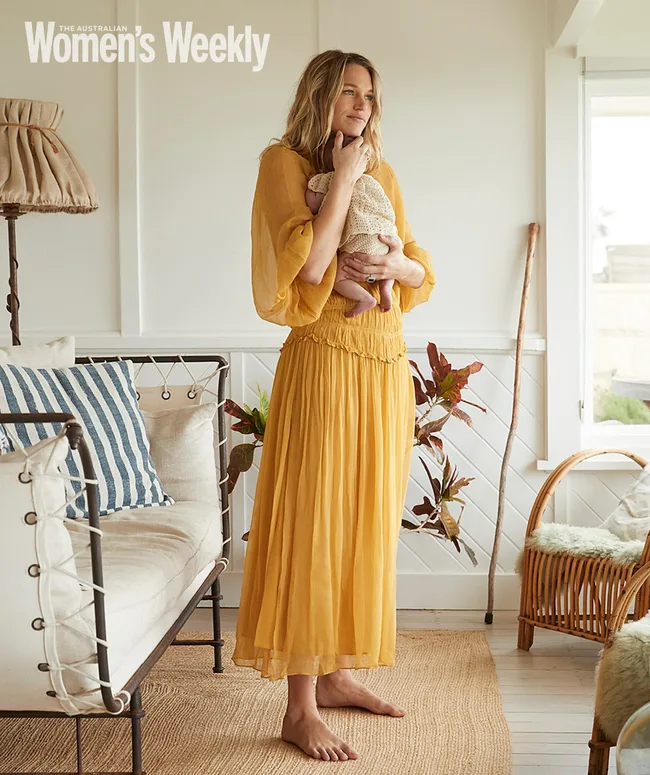 Ellidy Pullin stands barefoot in a living room, holding a baby, wearing a yellow dress.