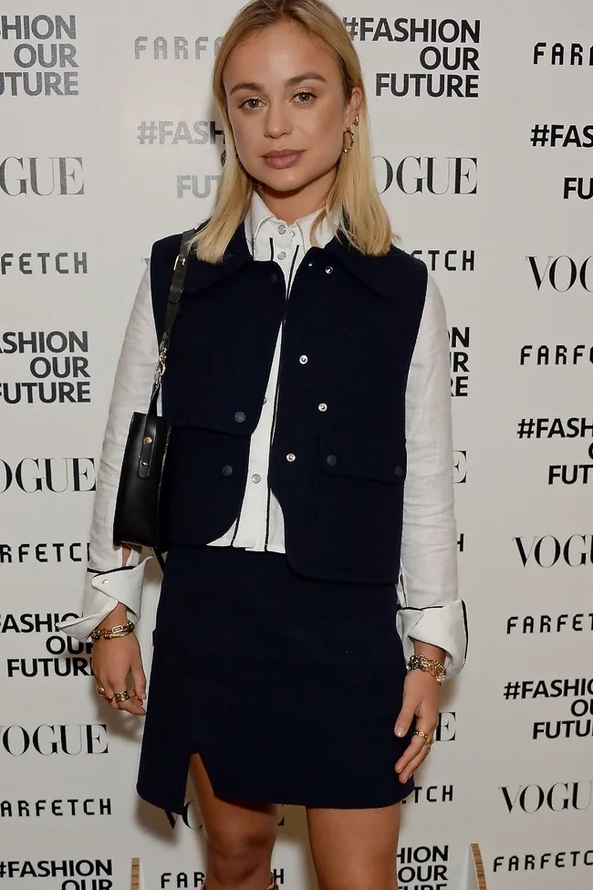 Young woman in a navy skirt and vest over a white shirt at a fashion event with Vogue and Farfetch logos.