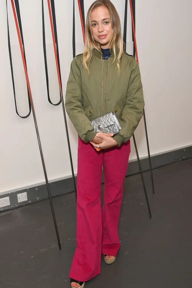 Woman in green jacket and pink pants holding a silver clutch, standing indoors with abstract art in the background.