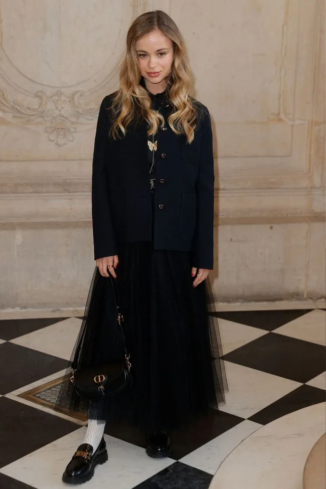 Young woman with long hair in black jacket and skirt, holding a small handbag, stands on checkerboard floor.
