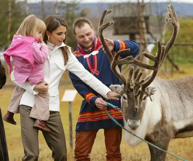 A woman and child pet a reindeer held by a man in traditional attire in an outdoor setting.