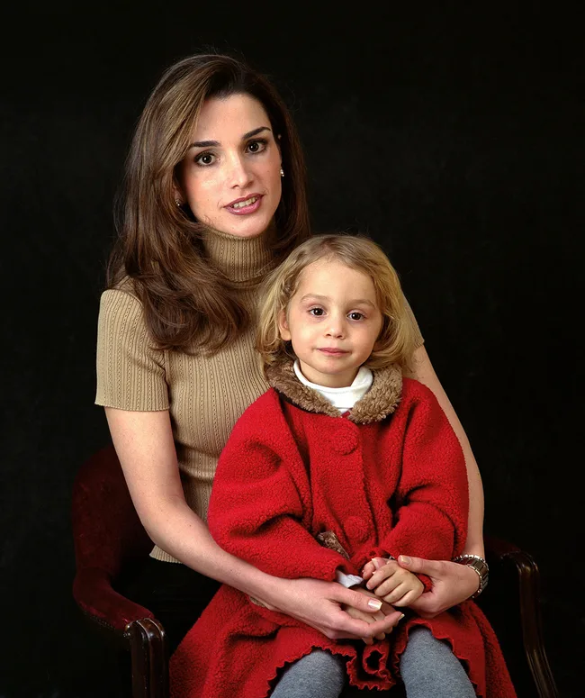 A woman in a beige sweater with long dark hair holds a young child in a red coat against a dark background.