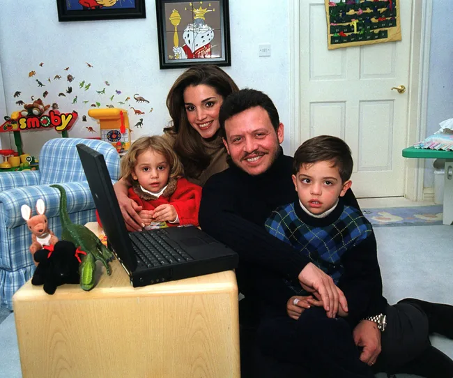 A family with two children poses together in a cozy, toy-filled room with a laptop on a small table.