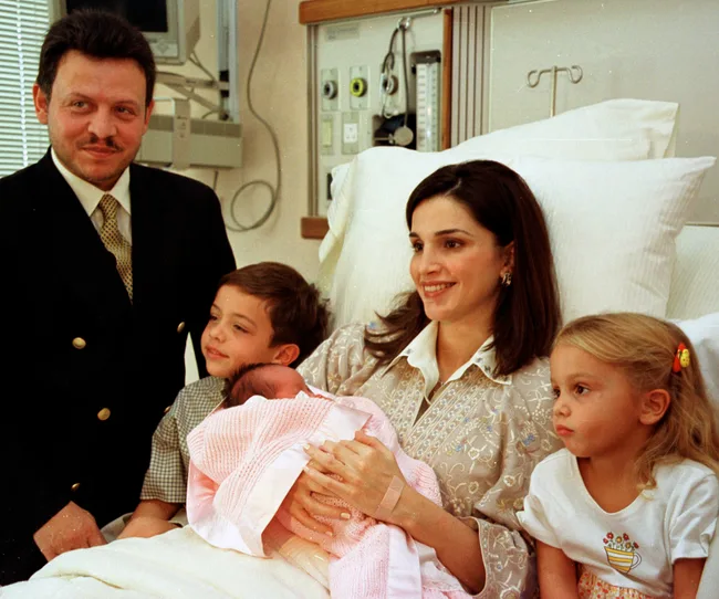 Family in hospital with mother holding a swaddled newborn, father standing, and two young children sitting beside her.