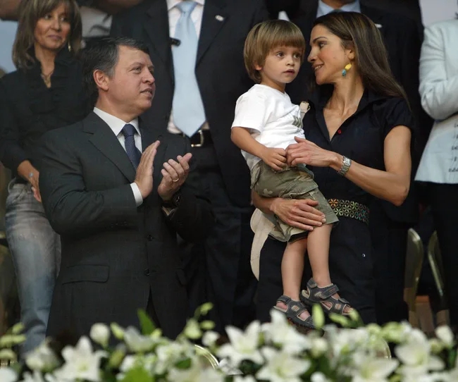 A man in a suit claps, while a woman in a dark dress holds a young child; they are surrounded by people and white flowers.