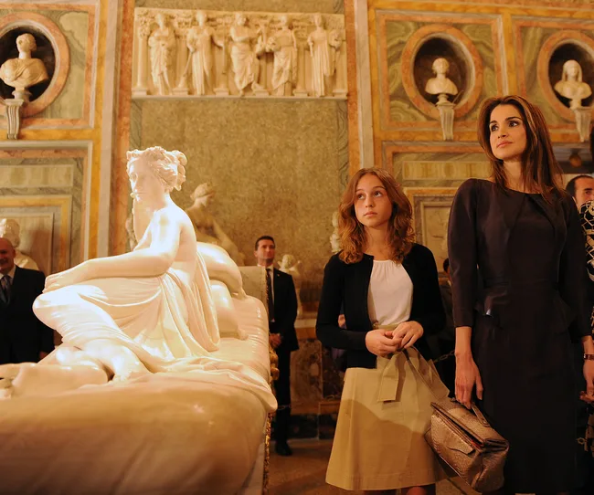 Two women observing a marble sculpture in an ornate gallery with historical carvings and decorative busts.