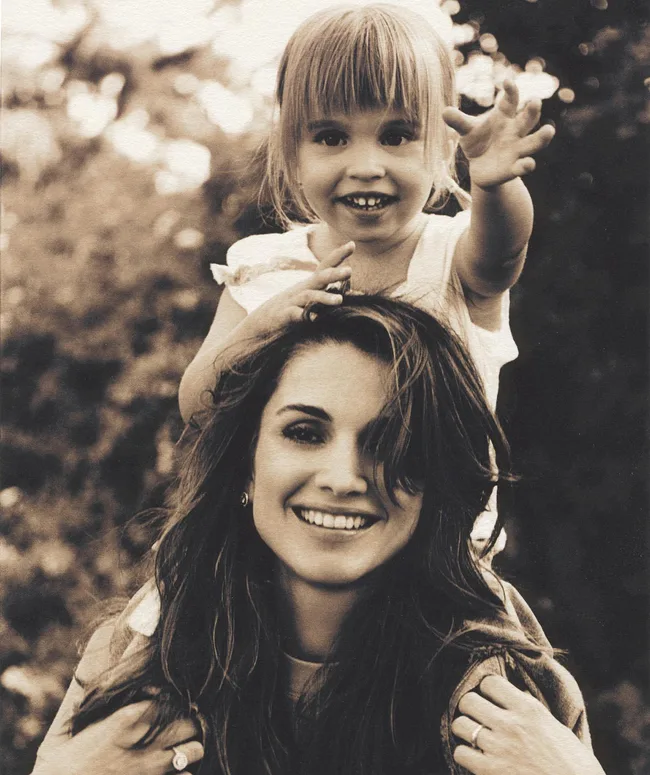 A woman smiling with a child on her shoulders, reaching out, in a sepia-toned outdoor setting.