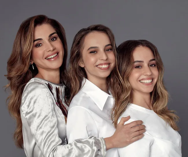 Three women smiling together against a gray background, wearing white outfits.