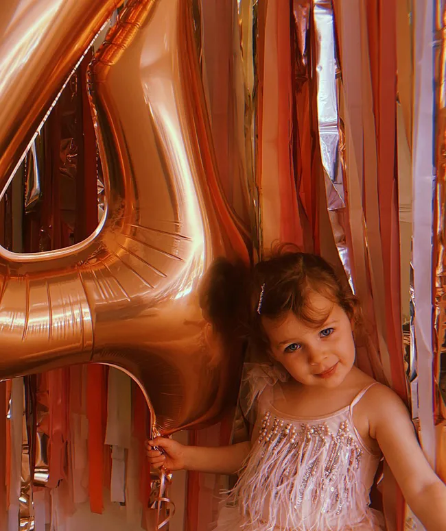 A child in a sparkly dress stands by a large gold balloon shaped as the number four, with colorful streamers in the background.