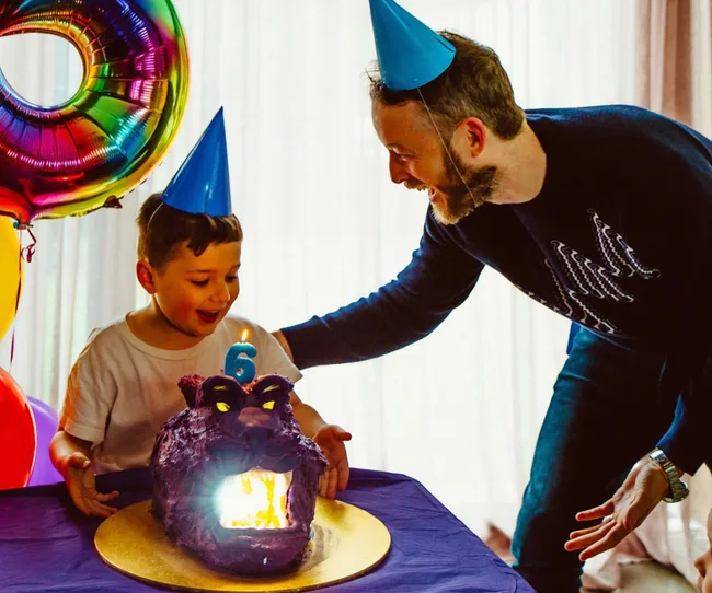 Boy with party hat smiles at a purple animal-shaped cake with a lit candle, next to a man at a birthday celebration.