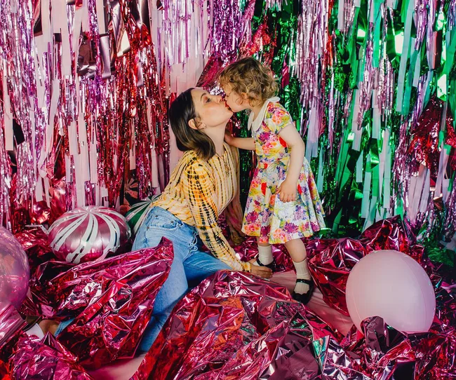 A woman and child kiss in a colorful confetti-filled room, surrounded by balloons and shiny decorations.