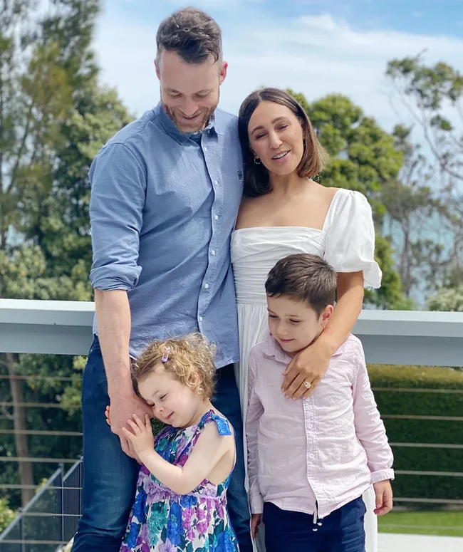 A family of four smiling outdoors, with lush greenery in the background.