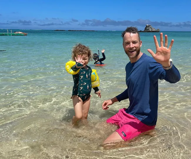 Man and child with floaties wave in clear beach water under a sunny sky.
