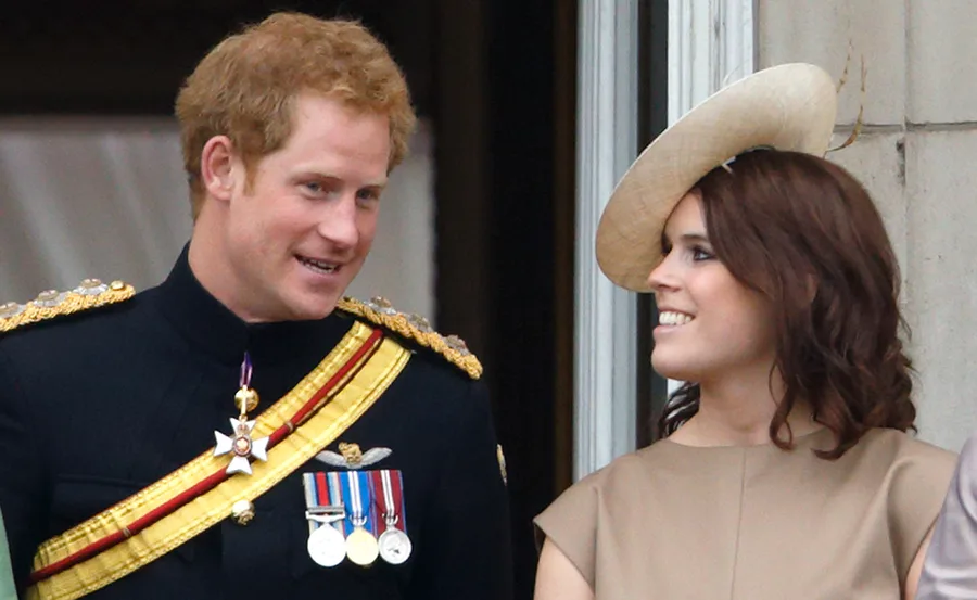 Prince Harry in a military uniform smiling and talking to Princess Eugenie, who is wearing a beige hat and dress.