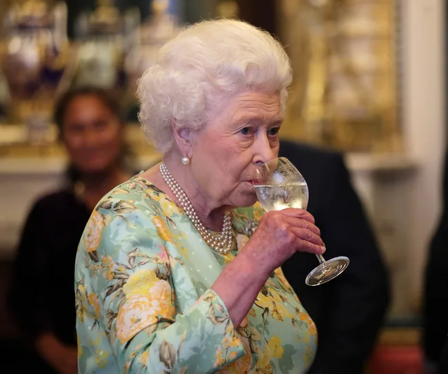 Elderly woman in floral dress drinks from a wine glass at a formal event.