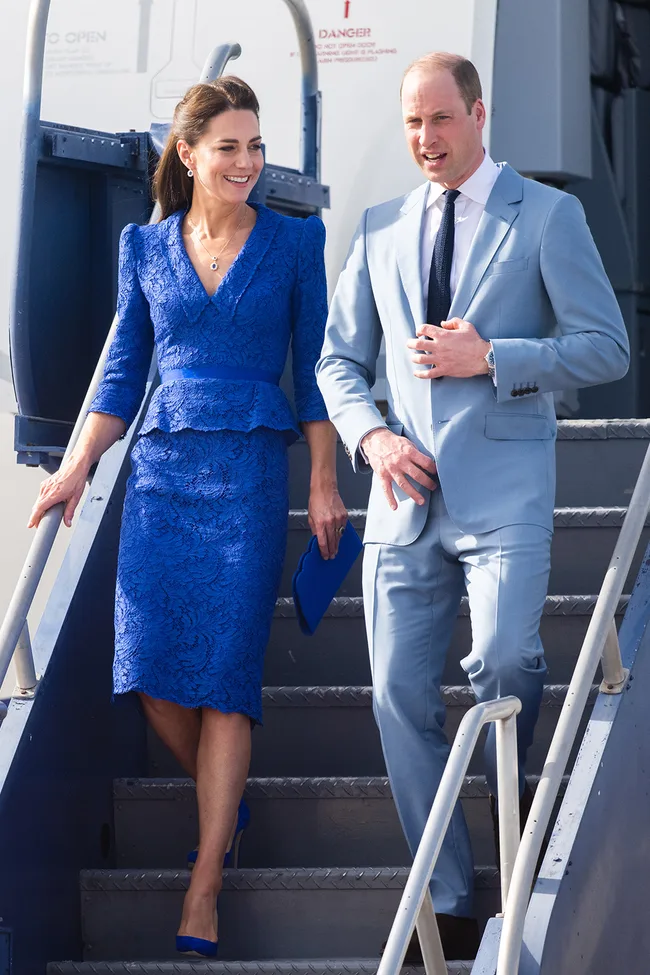 A woman in a blue lace dress and a man in a light blue suit descend airplane stairs, smiling.