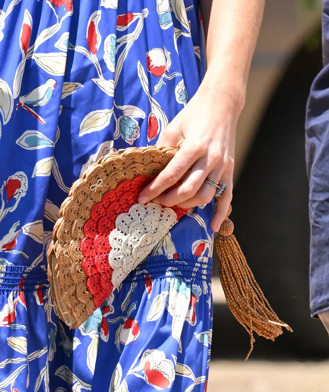 A hand holding a woven clutch with tassel against a floral blue dress.