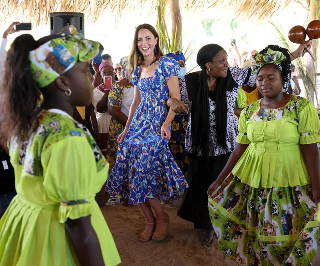 A woman in a blue dress dances with people in bright, festive clothing under a thatched roof.