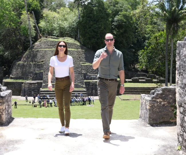 People walking near ancient stone ruins surrounded by trees and grass, with a group of tourists in the background.