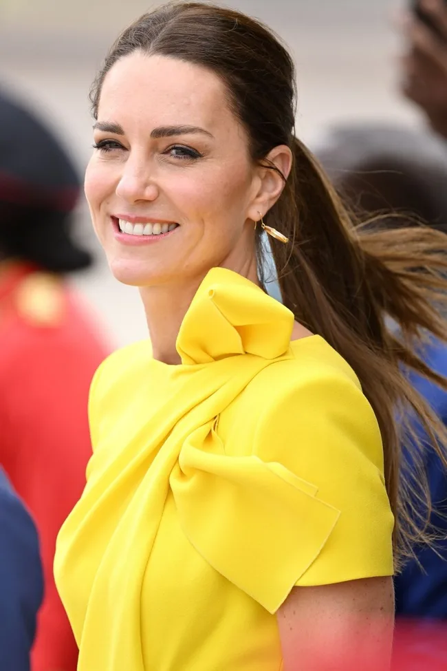 A woman with brown hair smiles, wearing a bright yellow dress with a bow detail on the shoulder.