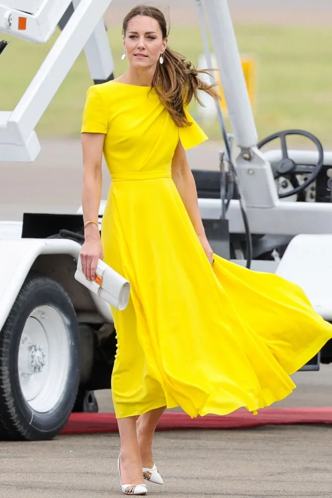 A woman in a flowing yellow dress walks outside near a vehicle, holding a small white clutch.