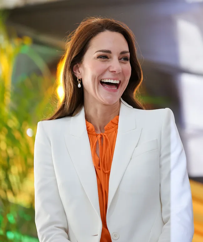A woman in a white blazer and orange blouse, smiling brightly with dangling earrings, standing in a well-lit area.