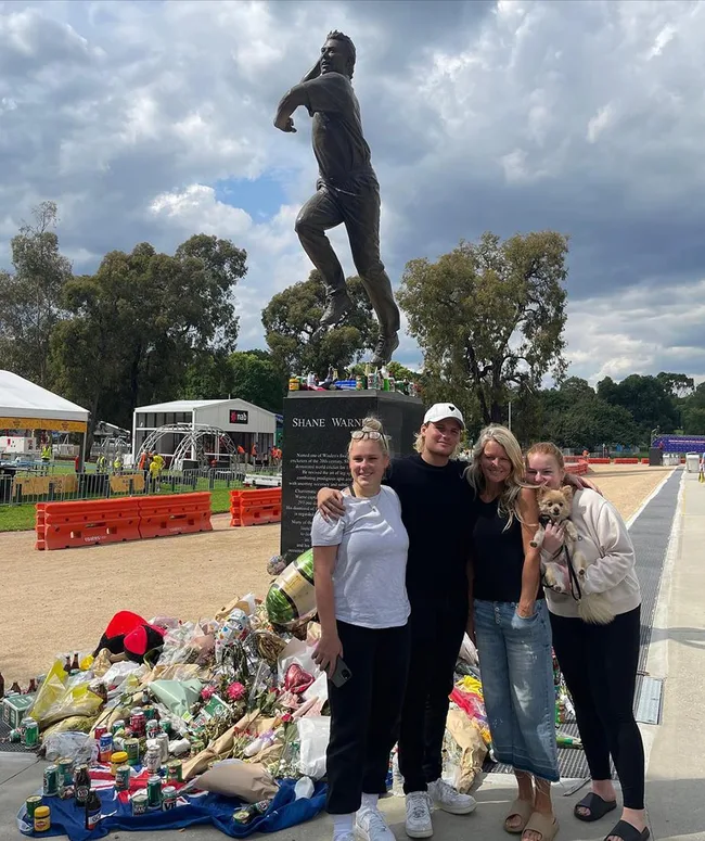Group standing beside Shane Warne's statue with flowers and tributes at the base.