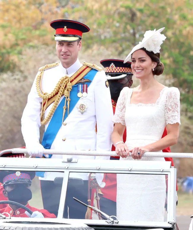 A man and woman in formal attire with military accents riding in an open vehicle, surrounded by others in uniform.