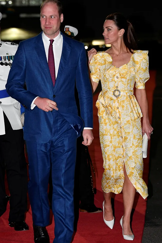 Man in blue suit and woman in yellow floral dress walk on a red carpet at night, flanked by a uniformed guard.